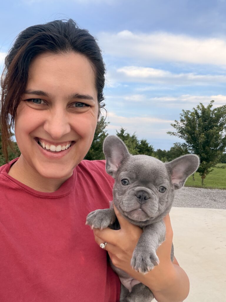 Smiling woman in a red shirt holding a gray French bulldog puppy outdoors, blue sky in the background.