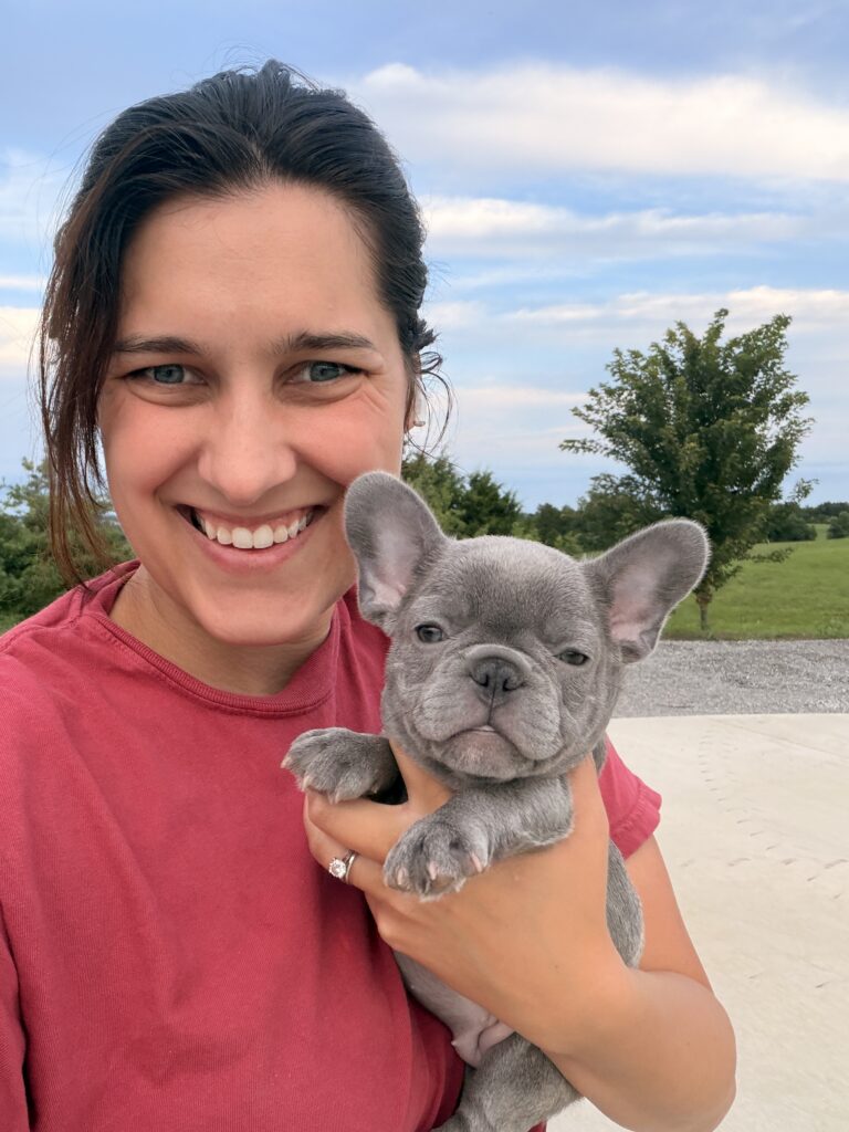 Smiling woman in a red shirt holding a gray French Bulldog puppy outdoors with a blue sky behind them